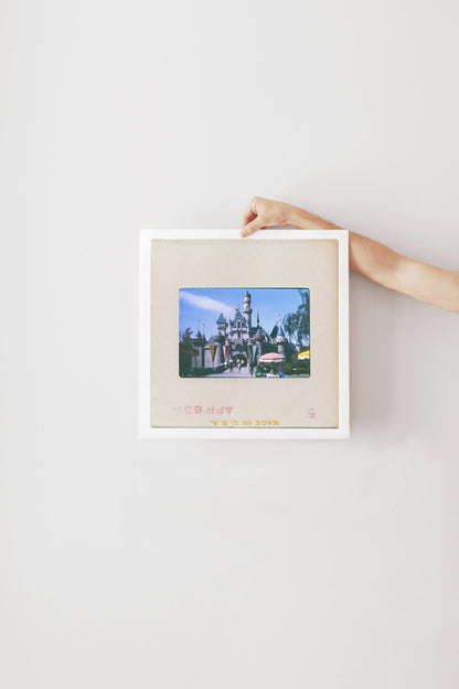A person's hand is holding a framed picture of a castle or palace, which appears to be a photograph of a Disney theme park or similar attraction.