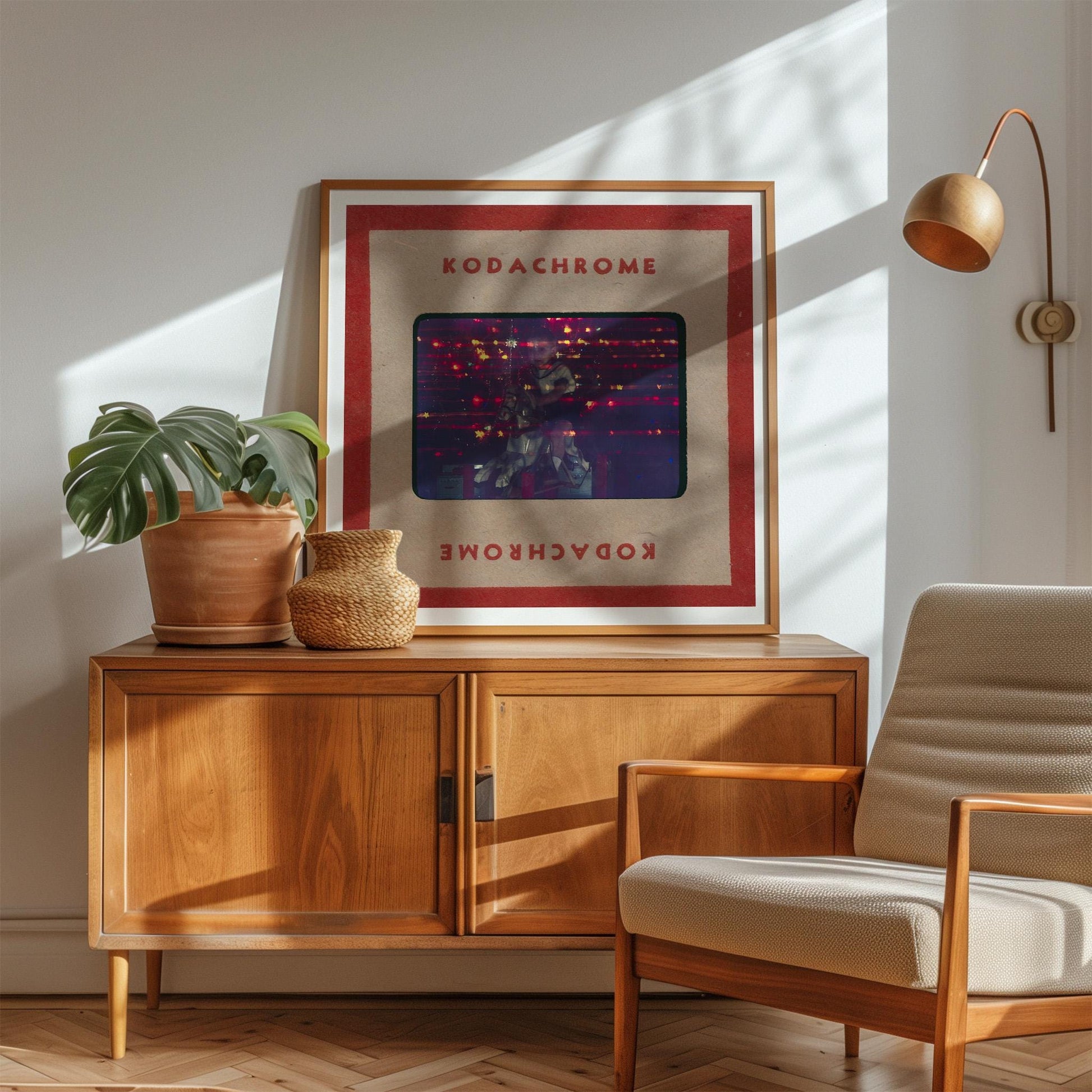 a wooden cabinet with a framed poster of Kodachrome's album cover on top, a potted plant, and a beige armchair in a sunlit room.