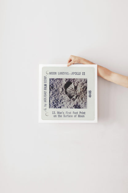 A person's hand is holding a framed photograph of a footprint on the surface of the Moon, which is labeled as "Man's First Foot Print on the Surface of Moon" and is part of the "Moon Landing - Apollo 11" series.