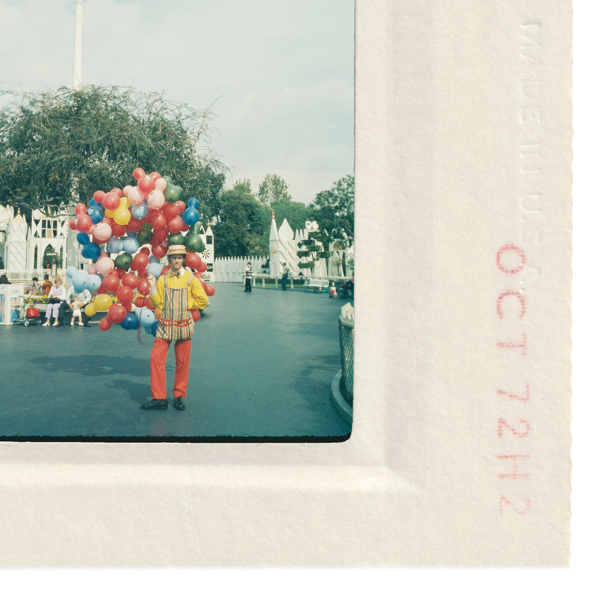 A clown holding a large bouquet of colorful balloons stands in a park-like setting, surrounded by trees and other people.