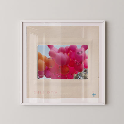 A framed photograph of a bunch of colorful balloons, including pink and orange balloons, floating in the air.