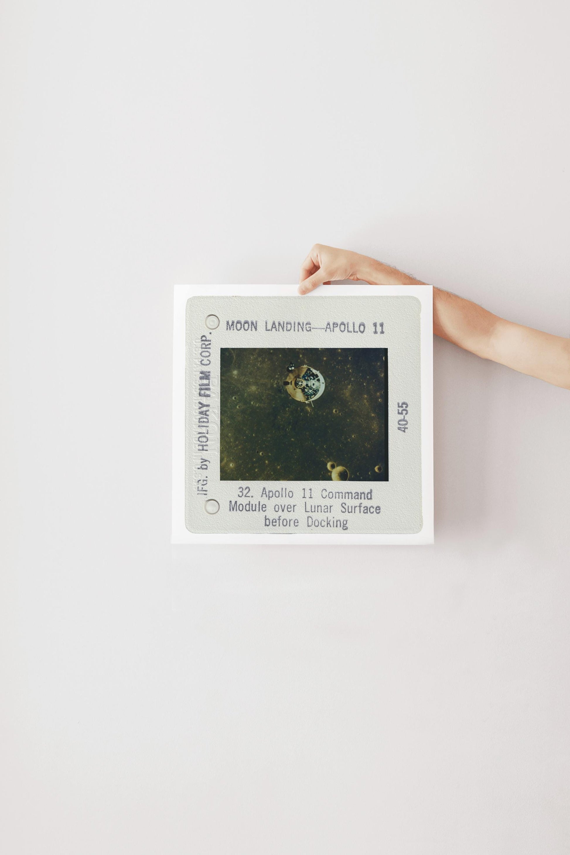 a person's hand holding a framed photograph of the Apollo 11 mission, which features the moon landing and the command module over the lunar surface.