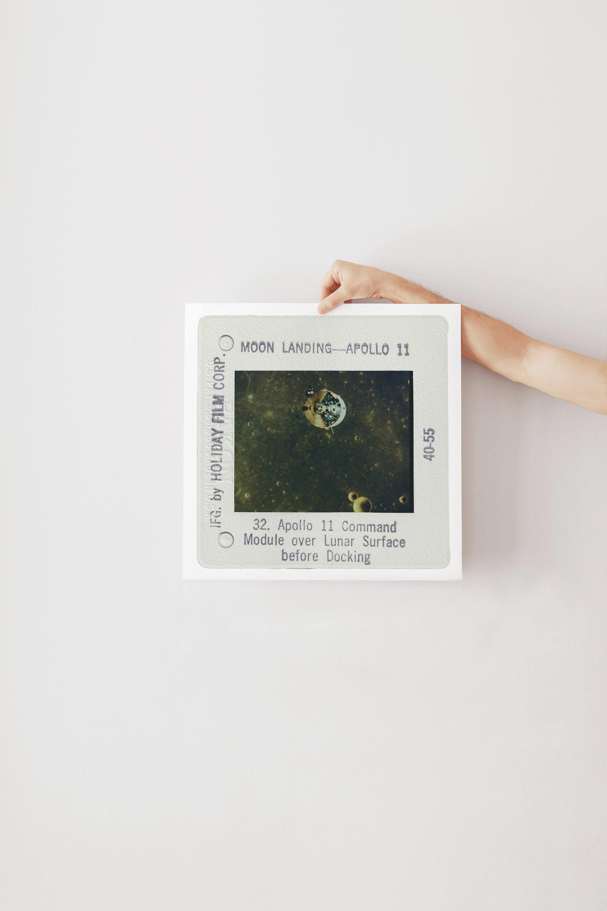 a person's hand holding a framed photograph of the Apollo 11 mission, which features the moon landing and the command module over the lunar surface.