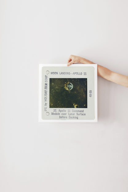 a person's hand holding a framed photograph of the Apollo 11 mission, which features the moon landing and the command module over the lunar surface.