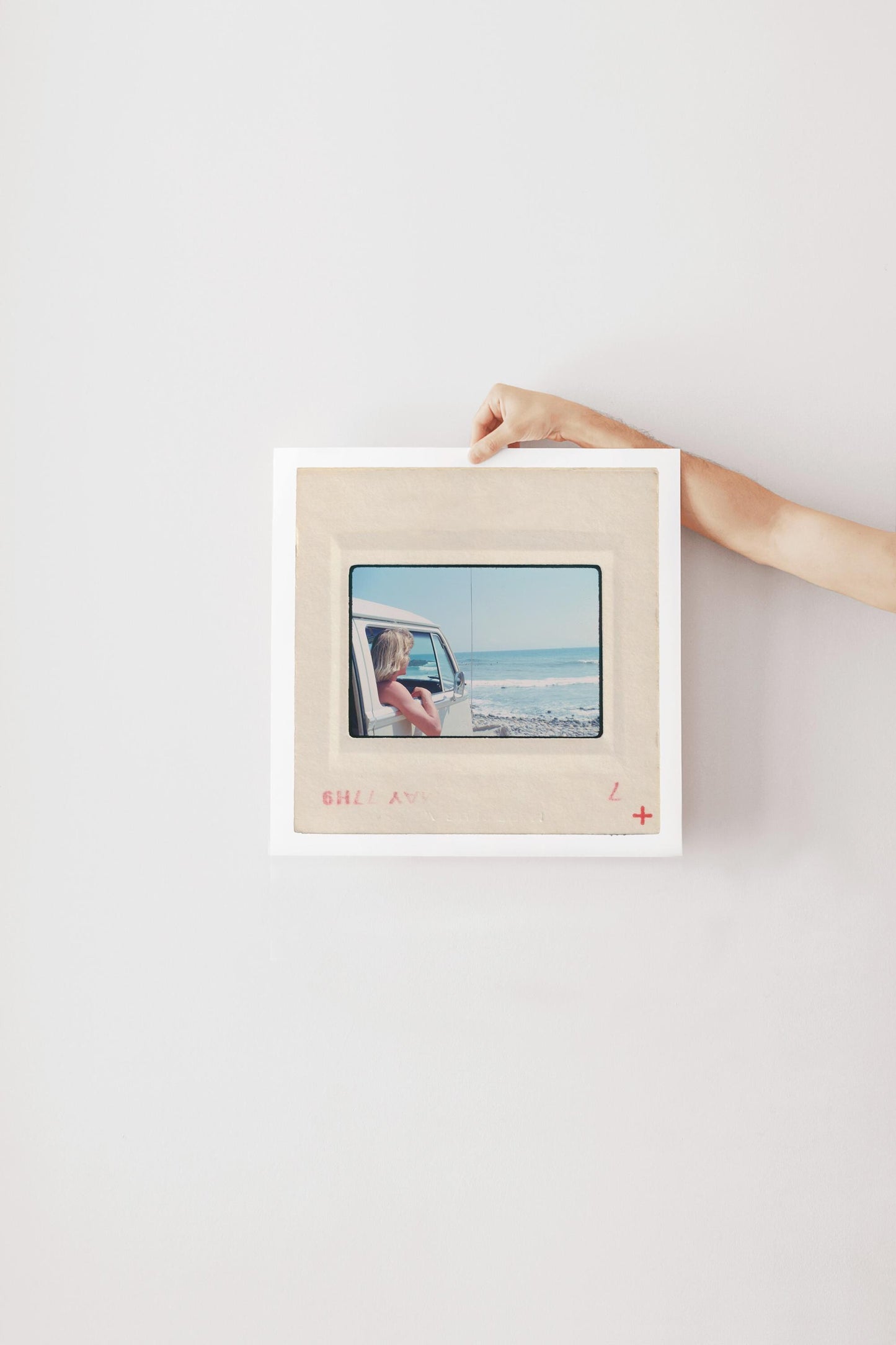 Vintage 1977 Kodachrome slide photograph of a blonde child leaning out the window of a white Volkswagen Microbus, looking toward the ocean along the California coast. Rocky shoreline and gentle surf visible in the background. Printed as a giclée fine art print on archival matte paper.