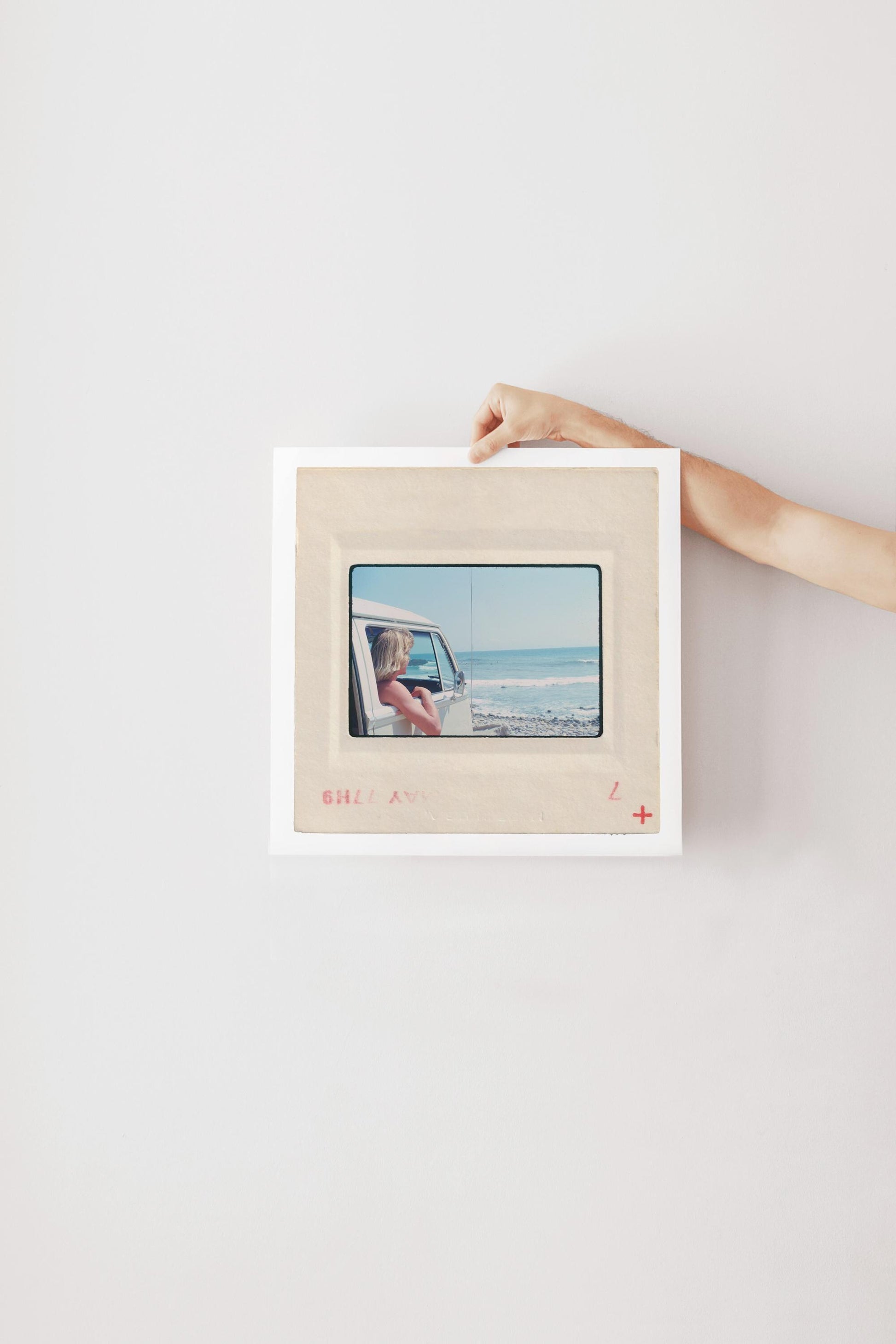 Vintage 1977 Kodachrome slide photograph of a blonde child leaning out the window of a white Volkswagen Microbus, looking toward the ocean along the California coast. Rocky shoreline and gentle surf visible in the background. Printed as a giclée fine art print on archival matte paper.