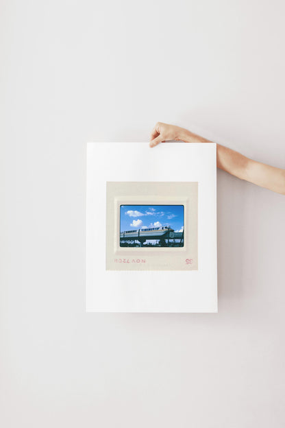 A person's hand is holding a framed picture of a train against a white wall.