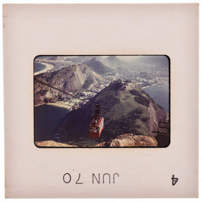 A red cable car is suspended above a mountainous landscape, with a city visible in the background.