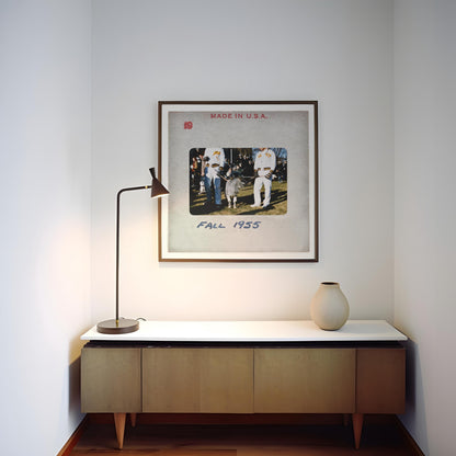 a framed photograph of people in a field, with the text "FALL 1955" visible. The photograph is mounted on a wall above a wooden cabinet or shelf, which has a lamp on top of it.