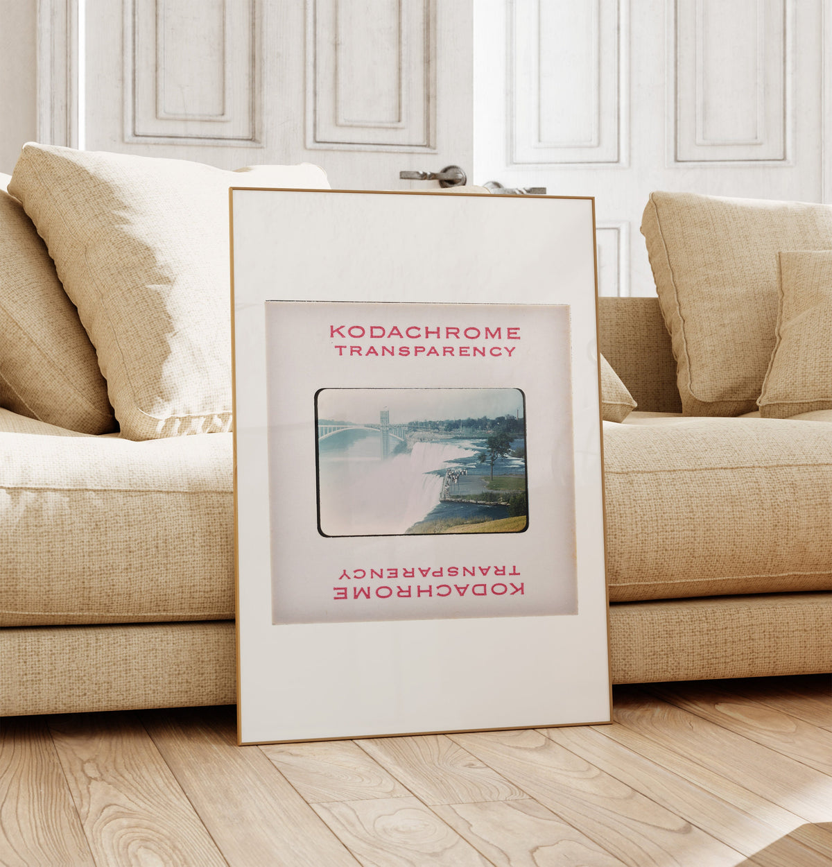 a framed poster of a waterfall, placed on a wooden floor in front of a beige sofa.