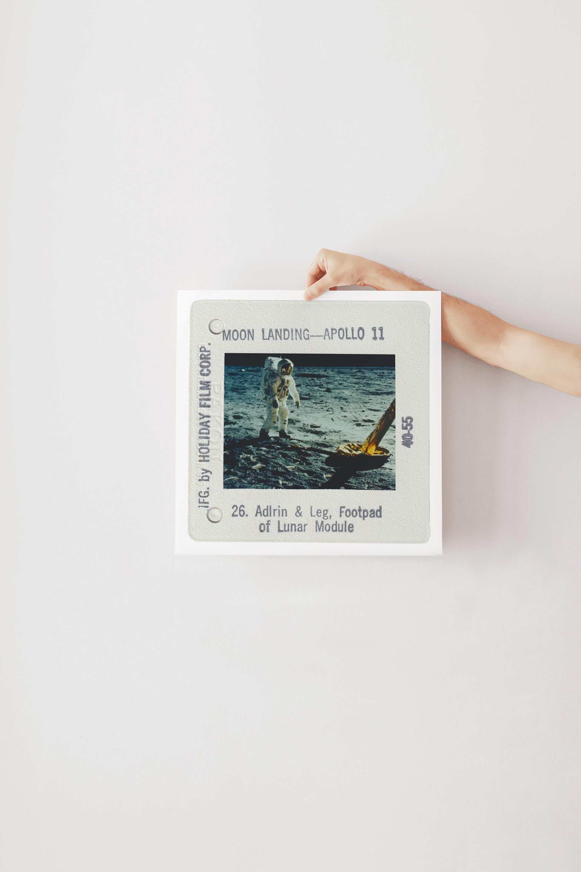 A person's hand is holding a framed photograph of an astronaut on the moon, with the text "Moon Landing - Apollo 11" and "26. Adirin & Leg, Footpad of Lunar Module" visible.