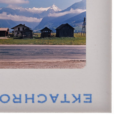 a rural landscape with a road, buildings, and mountains in the background.